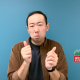 Paul ASL Video preview: A man with short dark hair stands against a teal blue background. He is wearing a dark t-shirt underneath an unbuttoned brown corduroy shirt. He has a neutral expression and looks toward the camera with his mouth slightly pursed. The framing is a medium shot capturing his head and shoulders in clear lighting.