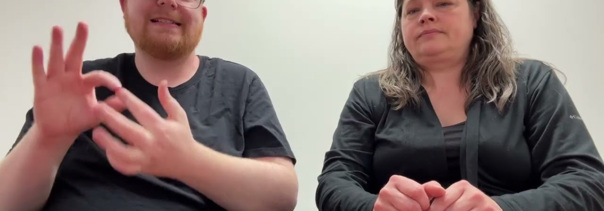 Youtube video preview, Two people are seated behind a brown wooden table against a plain white background. The man on the left has short reddish-blonde hair, a beard, and glasses, and he is wearing a black t-shirt. The woman on the right has long, dark, wavy hair and wears a black long-sleeved shirt. Both individuals look directly at the camera in a neutrally lit indoor environment.