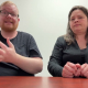 Youtube video preview, Two people are seated behind a brown wooden table against a plain white background. The man on the left has short reddish-blonde hair, a beard, and glasses, and he is wearing a black t-shirt. The woman on the right has long, dark, wavy hair and wears a black long-sleeved shirt. Both individuals look directly at the camera in a neutrally lit indoor environment.