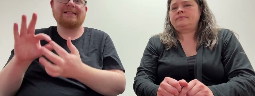 Youtube video preview, Two people are seated behind a brown wooden table against a plain white background. The man on the left has short reddish-blonde hair, a beard, and glasses, and he is wearing a black t-shirt. The woman on the right has long, dark, wavy hair and wears a black long-sleeved shirt. Both individuals look directly at the camera in a neutrally lit indoor environment.