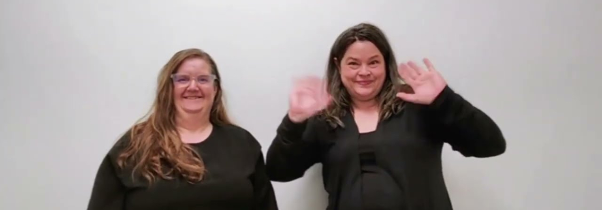 Two women stand side-by-side against a neutral, light-colored wall. The woman on the left has long, light brown hair and is wearing glasses with a black, short-sleeved top. The woman on the right has darker, wavy hair and is wearing a black cardigan over a matching shirt. Both are looking directly at the camera with neutral-to-slight smiles.