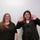 Two women stand side-by-side against a neutral, light-colored wall. The woman on the left has long, light brown hair and is wearing glasses with a black, short-sleeved top. The woman on the right has darker, wavy hair and is wearing a black cardigan over a matching shirt. Both are looking directly at the camera with neutral-to-slight smiles.