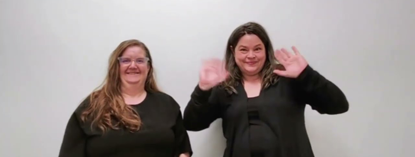 Two women stand side-by-side against a neutral, light-colored wall. The woman on the left has long, light brown hair and is wearing glasses with a black, short-sleeved top. The woman on the right has darker, wavy hair and is wearing a black cardigan over a matching shirt. Both are looking directly at the camera with neutral-to-slight smiles.